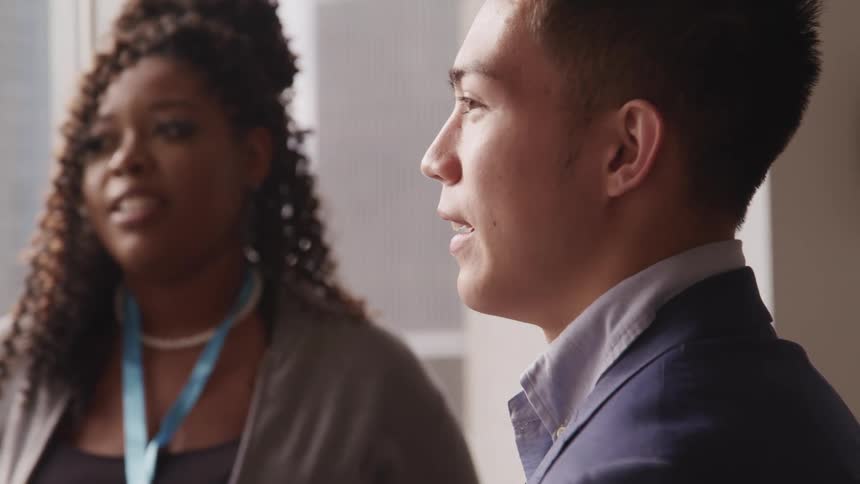 A young man and young woman smile and react to something they are told during a workplace conversation