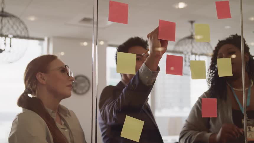 A man writes on a sticky note attached to a glass wall as his female colleagues watch and speak to him
