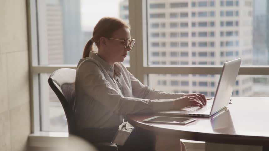 Female concentrates as she sits at round table next to window overlooking the city while typing on the keyboard of a laptop