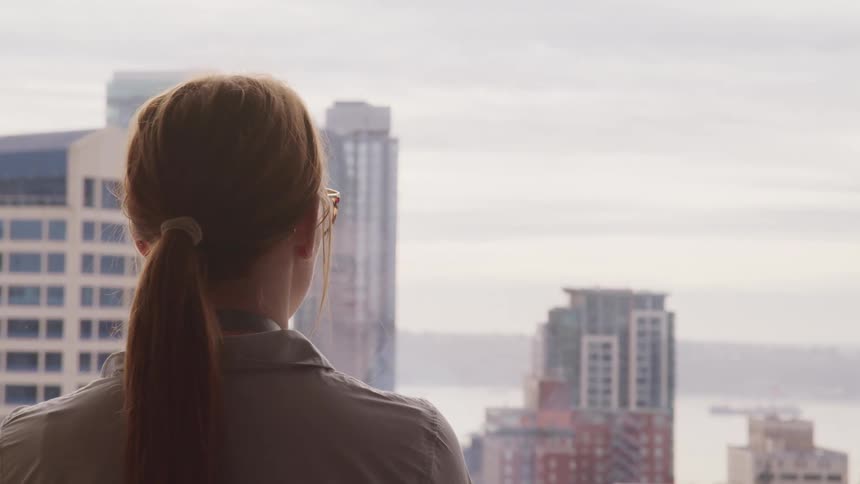 A woman with glasses standing in front of a window with buildings the sky and clouds in the background