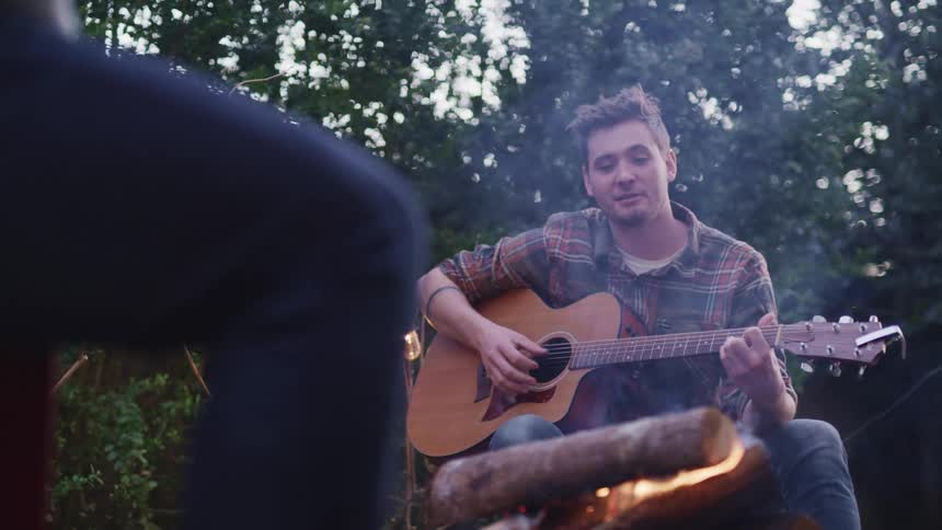 Two people sit around a campfire while one plays a guitar and sings