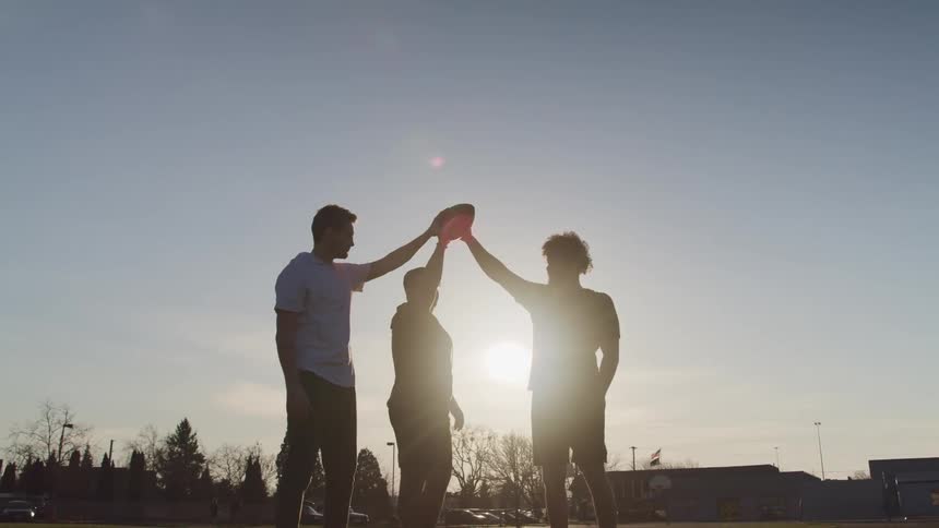 Silhouette of three friends fist bumping around a football in a field at sunset