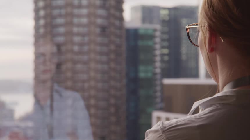 A woman crosses her arms as she looks out of her office window at skyscrapers downtown