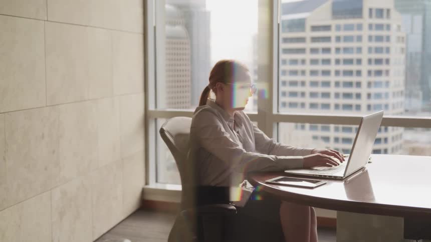 Woman sits in conference room typing on laptop next to large window showing city skyscrapers