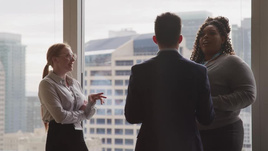 Three business professionals stand in an office beside large windows having a discussion