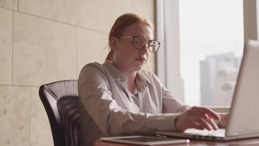 Businesswoman with red hair a shirt and glasses sits at a table and uses a laptop in the office