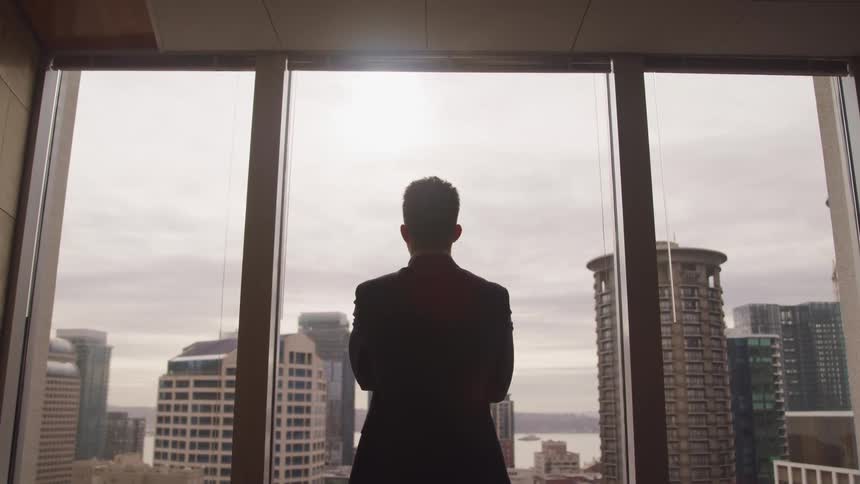 A man looks out his office window at the high rises across the street