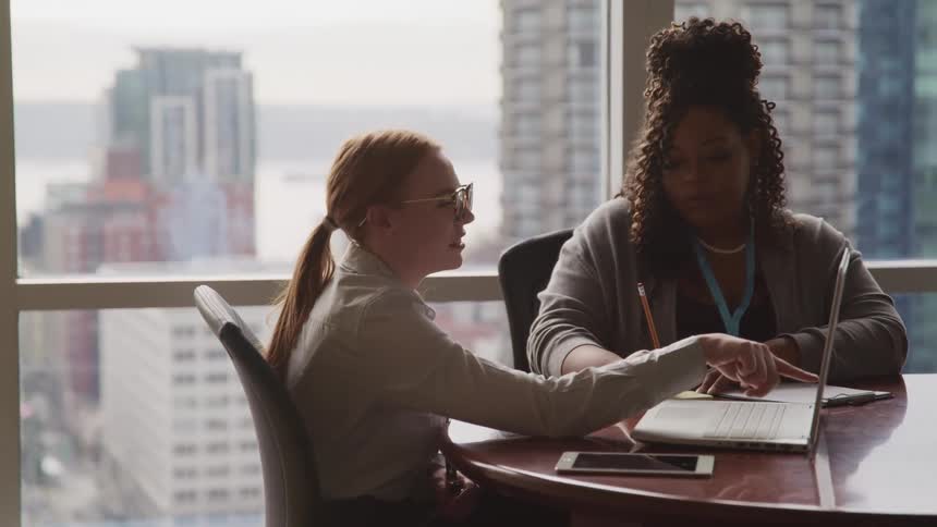 Boss points at a laptop while dictating to her secretary in an office in the city