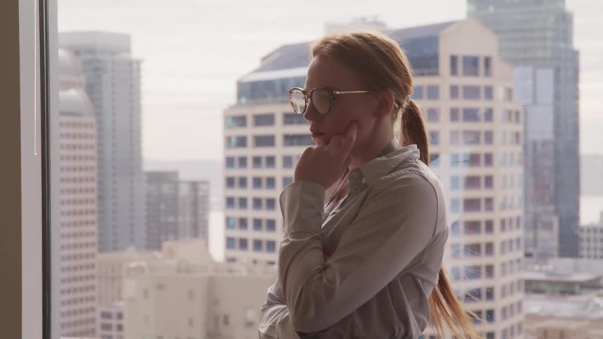 Businesswoman with red hair and glasses looks out the window in the office