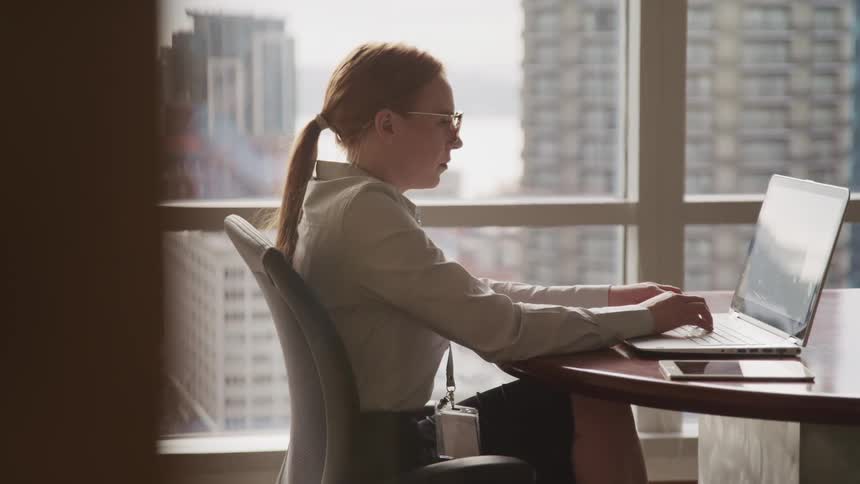A woman sits at a table in an office typing on a laptop