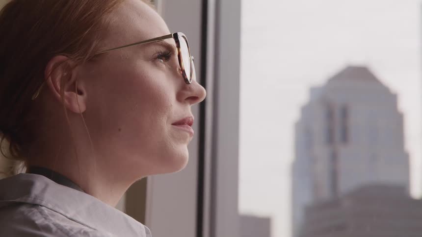 A woman wearing glasses looks thoughtfully out a large window that provides a view of several very tall buildings