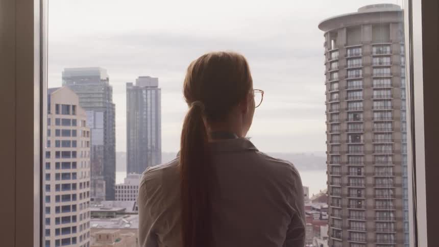 A woman looks out a window at the high rise buildings across the street from her