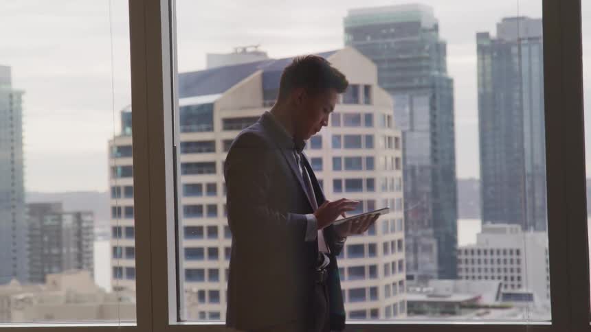 Businessman in a suit uses a tablet next to office windows with tall buildings in the background