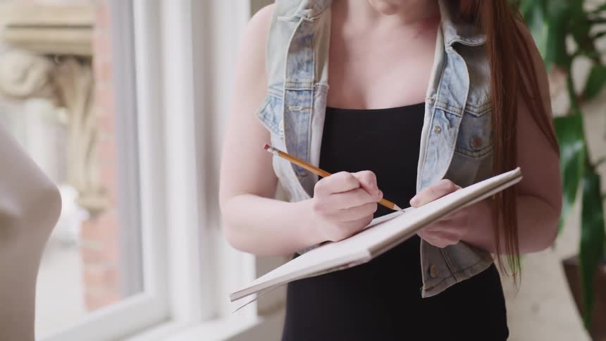 A woman standing next to a window and holding a notepad and pencil