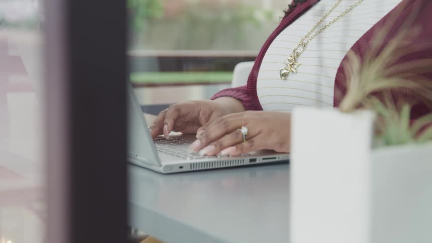 Black chubby woman with a necklace and a ring sits at a desk and uses a grey laptop
