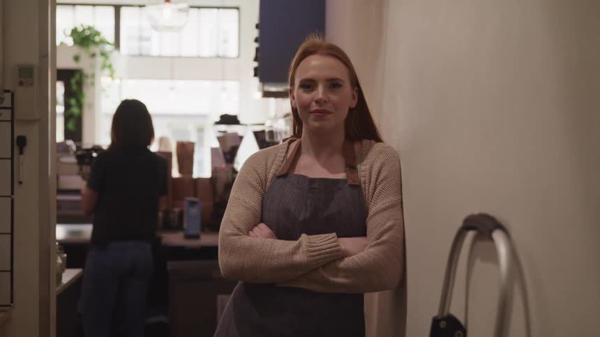 A barista poses with her arms crossed in the back hallway of a coffee shop as another employee makes drinks behind her