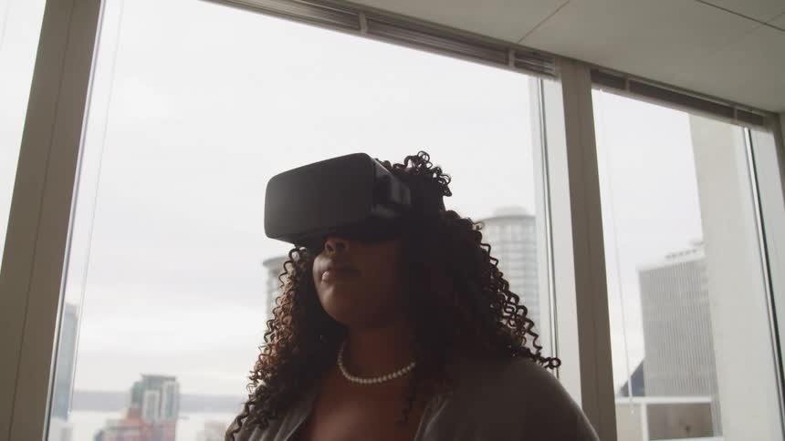 A woman in formal clothing stands in an office while wearing a virtual reality headset