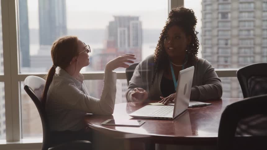 Two female coworkers sit at a table discussing something