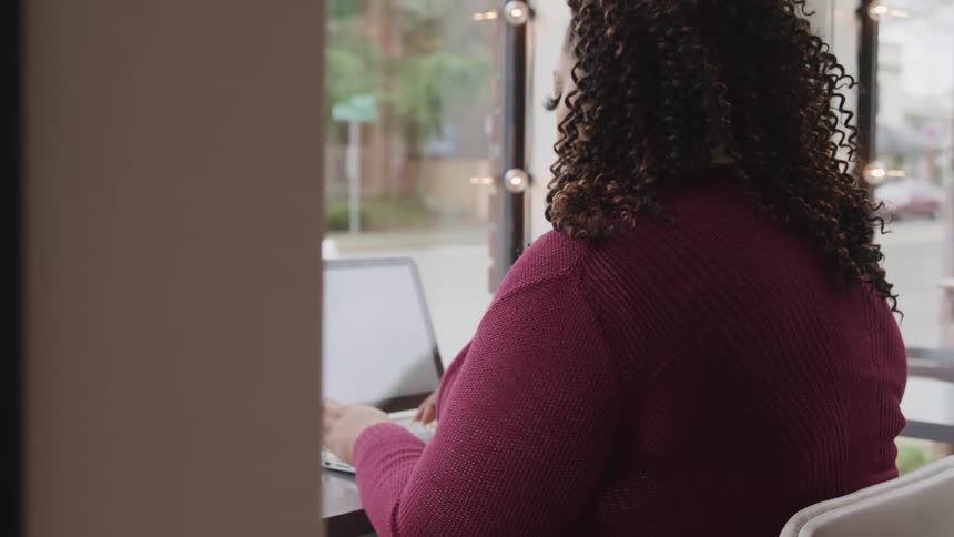 A black woman in a sweater sits at a desk in front of window while typing on a laptop