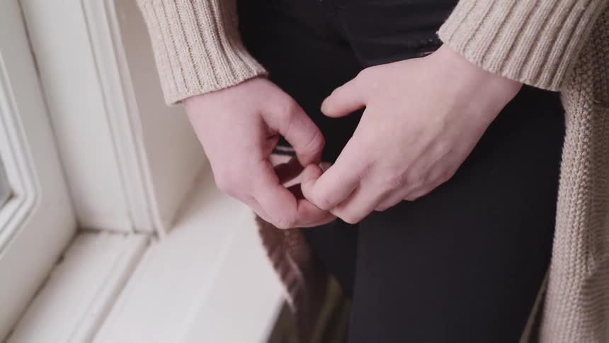 Woman wearing a sweater and ripped jeans fidgets her hands standing near window