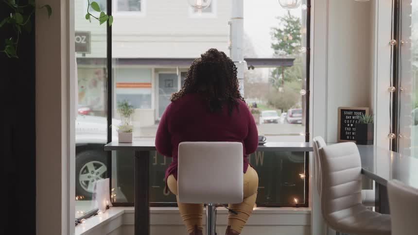 A woman sits alone at a cafe table looking out of the large window in front of her