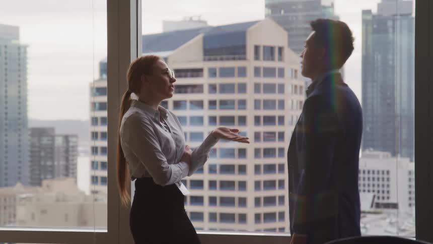 Businesswoman with red hair glasses and businessman with a suit talk to each other by the office window in the office