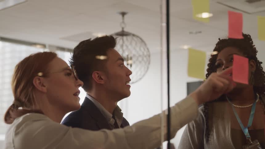 A man and two women looking at a sticky note on a glass with a window in the background