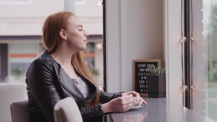 A woman sits at a window side table in a cafe with a small cup of coffee