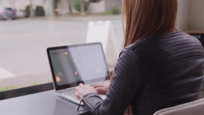 A woman types on her laptop in front of a large window with views of her neighborhood