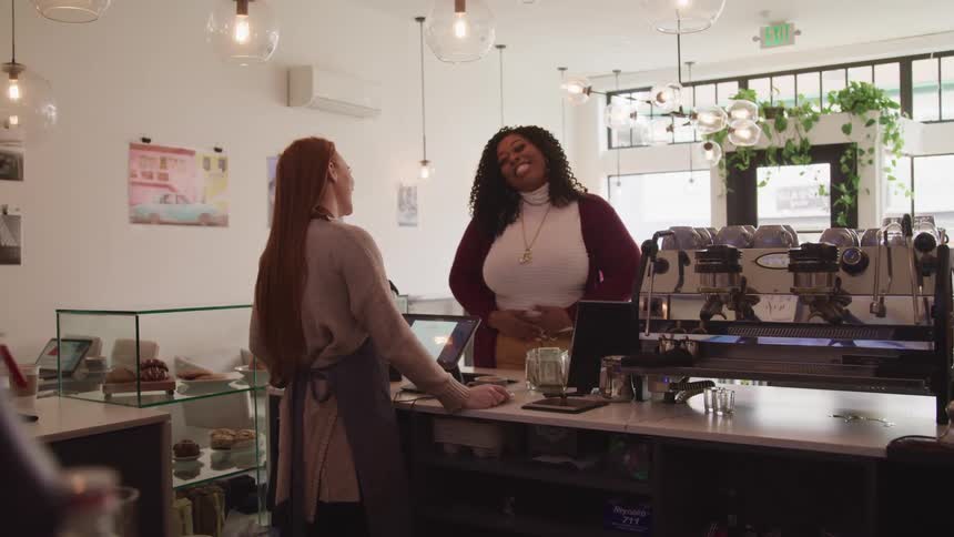 Counter worker and customer laugh and talk over counter near coffee machines and baked good display case