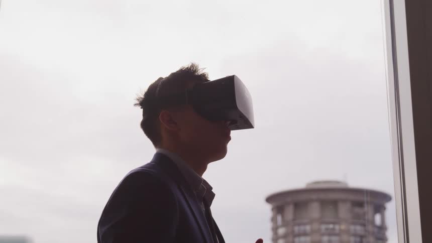A man stands wearing vr glasses with his fingers and hands moving with a cloudy city sky behind him