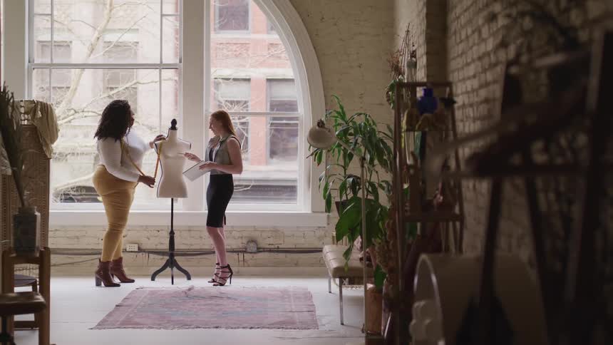 Two women discuss a design while using a measuring tape on a dress form in a business