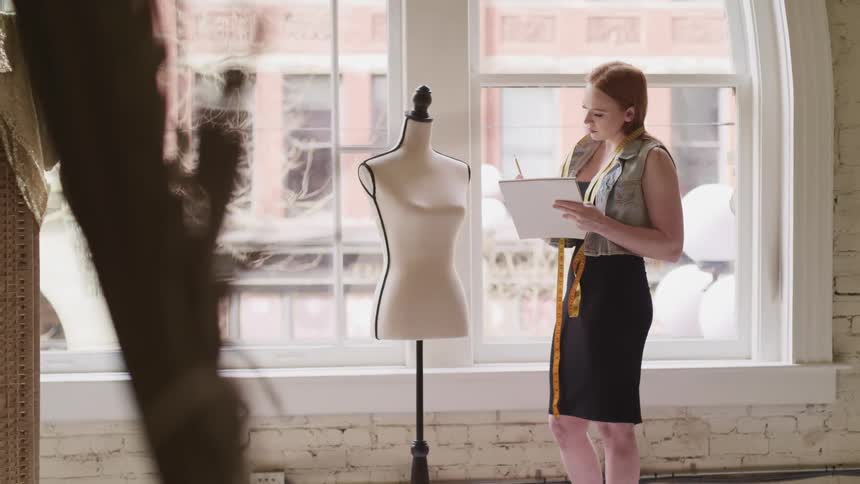 Fashion designer takes note on a notebook with a pencil in front of a mannequin in a studio