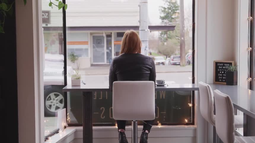 A woman sits at a table in a cafe staring out the large window in front of her towards the street