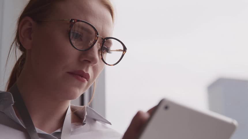 A woman with glasses and a tablet next to a window with a building in the background