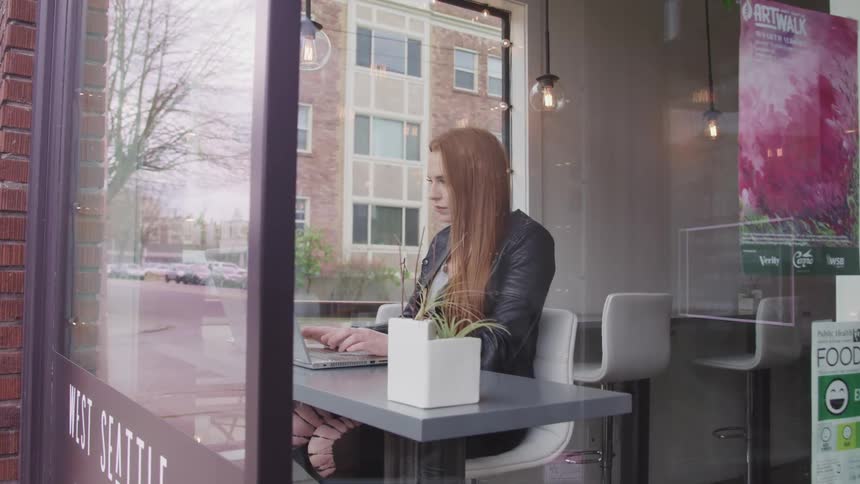 Woman with red hair and a black leather jacket sitting at the table uses a laptop in a cafe
