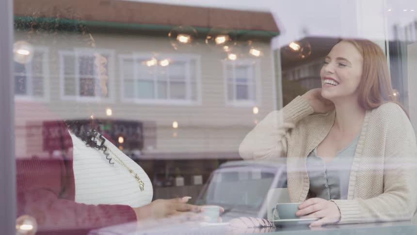 White woman with red hair and chubby black woman sitting at a table talk to each other and laugh together in a cafe
