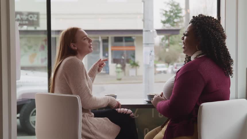 Two woman sit in a cafe at a window side table talking and laughing with small coffee mugs on their table