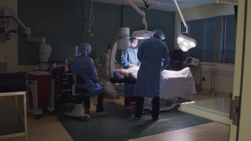 A pair of surgeons perform surgery on an unconscious patient as a nurse monitors the patients vitals