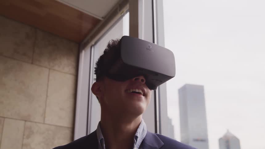A man stands next to a window in a city and smiles while using a virtual reality headset