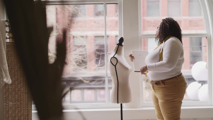 A clothing designer measures a mannequin and then looks at her paper