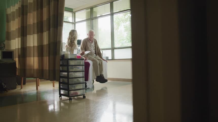 A female medical professional sits with an elderly male patient in a hospital room