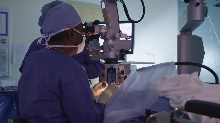 A surgeon uses a surgical microscope to perform a surgery alongside another doctor