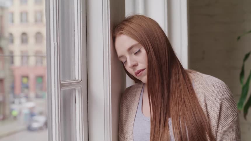 Woman with red hair leans against a wall next to the windows and looks down