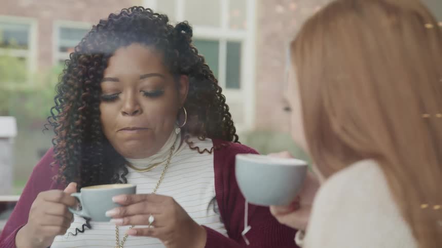 Black woman with curly hair and white woman with red hair sit at a table and drink coffee and tea in a cafe