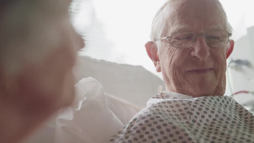 An elderly man in a medical gown sits upright in bed and smiles at his female companion