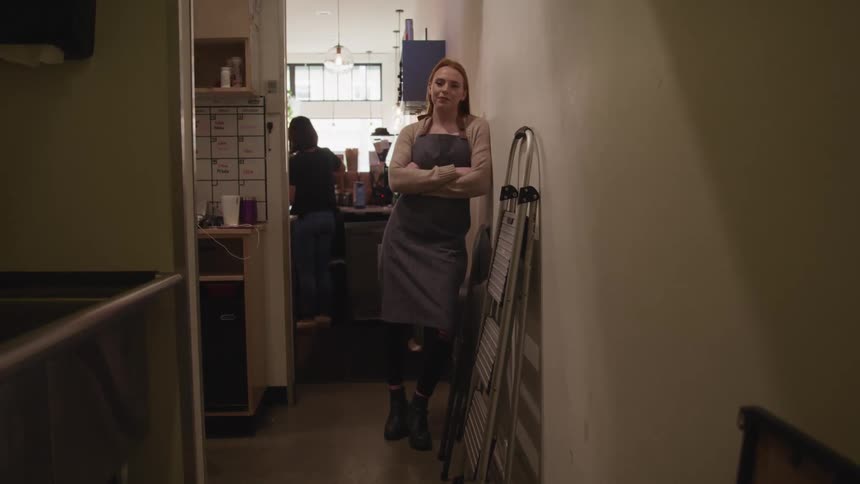 A woman in apron leans against the wall in the backroom of a cafe and looks into the camera as it approaches