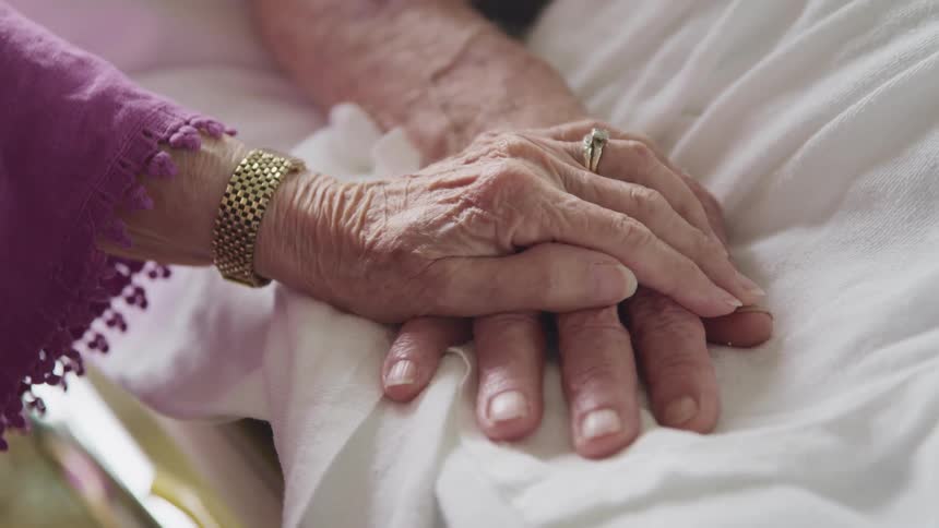An elderly woman rests her hand on her husbands as he lies in a hospital bed