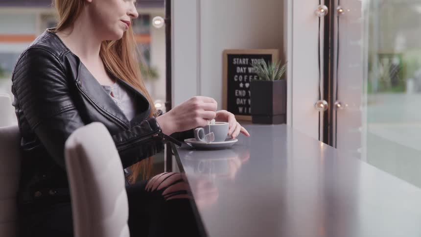 A woman sits at table in front of a window in a cafe and steeps her tea