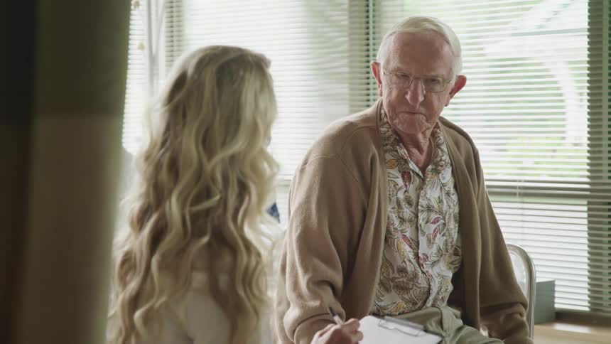 A female doctor sits beside an elderly man in a hospital room or clinic room